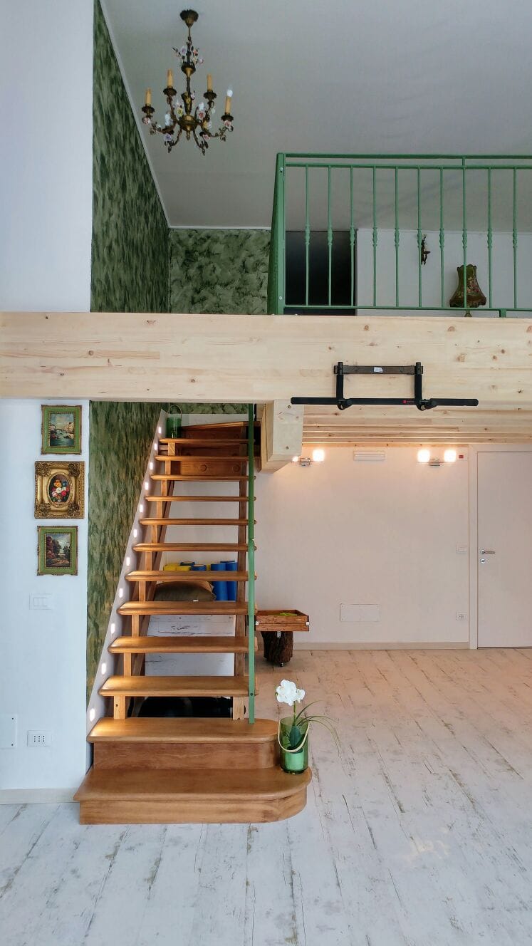 Ambiente a doppia altezza con soppalco, scala in legno e parete decorativa verde.